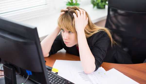 A woman with blond hair wearing a black shirt leans her elbows on her desk and holds her head in her hands as she stares in frustration at a computer screen