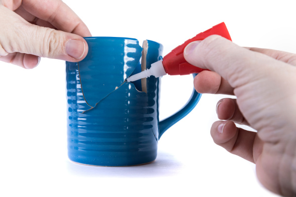 In front of a white background a pair of hands uses a red superglue tube to repair a blue coffee mug.