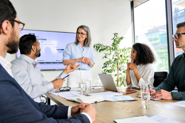 A diverse group of people sit around a conference table while a woman gives a presentation.