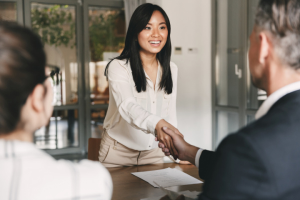 A woman reaches across a desk to shake the hand of the man sitting behind it. A resume is on the desk between them. Another woman, facing away from the camera, is in the foreground behind the desk.