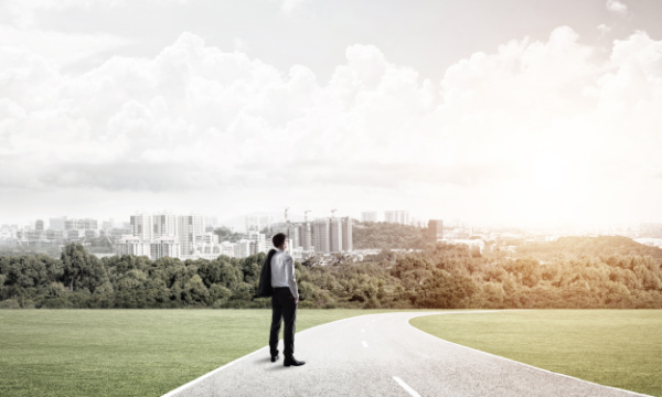 A man with his suit jacket slung over his left shoulder stands on a road looking at a city in the near distance. On the left side of the image, the city is grayer. On the right side it's more brightly lit.