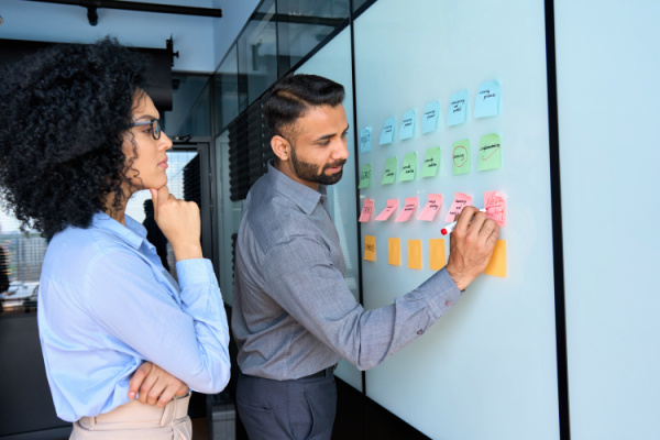 A woman looks thoughtful as a man writes on colorful sticky notes posted on a whiteboard.