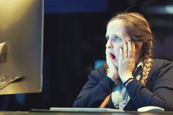 A woman with a shocked expression on her face stares at a computer screen. Her hands are on her cheeks.