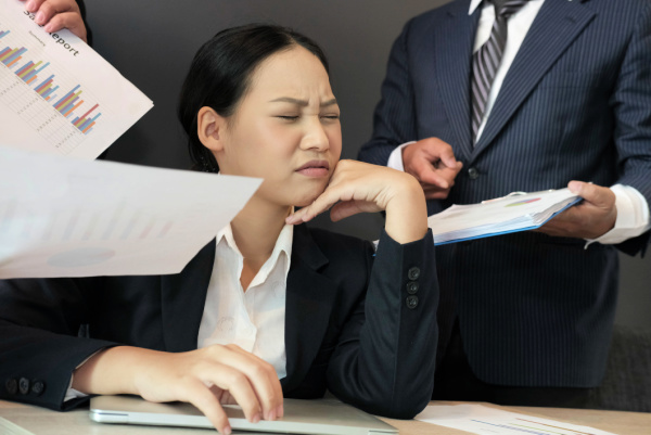 a woman in a black suit at a desk is overwhelmed with people approaching her for help.