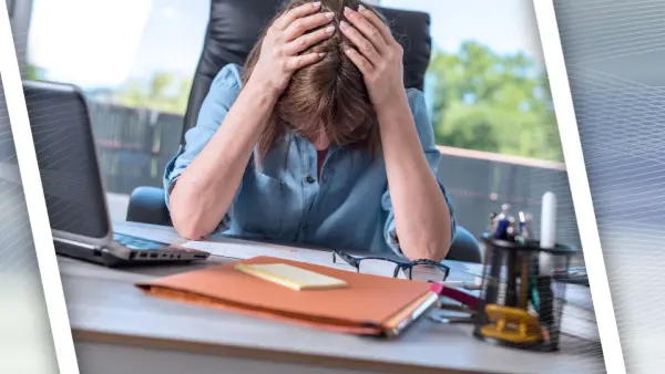 A woman sits at a desk with her head in her hands, surrounded by folders, papers, and office supplies, suggesting workplace stress and overwhelm.