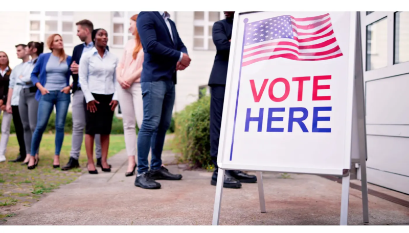 A 'Vote Here' sign with an American flag stands outside a polling location while a line of voters waits nearby — representing the election-day preparation and worker safety planning covered in this post.